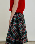 A person wears Aquarius Cocktail's Le Cargo Skirt—a long, black plaid piece with red, green, blue, and yellow stripes—paired with a red short-sleeve top. Their tattooed right arm rests at their side. Skirt is made in Los Angeles; background is light and plain.