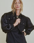 A woman with wavy blonde hair models the Ava Cropped Jacket Organza Sleeve by Aquarius Cocktail, featuring silk organza sleeves and large front pockets, styled with a chunky necklace against a plain light background.