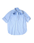 The "Dear Polly Shirting Bow Blouse" by Aquarius Cocktail is a light blue, short-sleeved blouse made from Japanese cotton shirting, adorned with thin vertical stripes. It boasts a large bow tied at the neck and has a loose, casual fit. The menswear-inspired detailing includes buttons down the front and a curved hemline for an elegant finish.