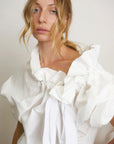 A woman with wavy blonde hair wears the Beautiful Mess Poet Blouse by Aquarius Cocktail, featuring sculptural ruffles and white ribbon accents, standing against a plain light background.