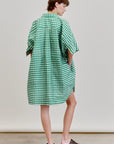 A person faces away from the camera, wearing the Aquarius Cocktail Ging Bellini Dress—an oversized green and white gingham shirt dress—and black woven slide sandals, set against a plain light background.