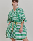 A model in the Aquarius Cocktail Ging Bellini Dress—an oversized green and white gingham shirt dress with a gold chain belt—stands before a plain light background, confidently gazing at the camera.