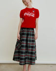 A woman stands against a plain background wearing a red Coca-Cola T-shirt, shiny gold flats, and the Le Cargo Skirt by Aquarius Cocktail—a long Japanese cotton poplin plaid skirt with red and green lines. She gazes at the camera, head slightly tilted.