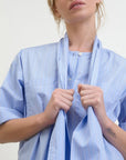 A person wearing the Dear Polly Shirting Bow Blouse by Aquarius Cocktail, crafted from light blue, pinstriped Japanese cotton shirting, stands against a neutral background. They are adjusting the matching fabric bow around their neck, with part of a tattoo visible on their forearm.