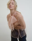 A person with wavy blonde hair leans forward, wearing the Aquarius Cocktail Dear Polly Bow Blouse—a sheer, peach oversized fit with a large bow at the collar. Against a plain white backdrop, they complete their look with dark pants.