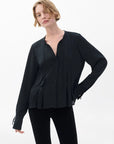 A person with short, wavy blonde hair wears the Rag & Bone arden pleated blouse in black with long sleeves and black pants, standing against a plain light background with one hand on their hip.