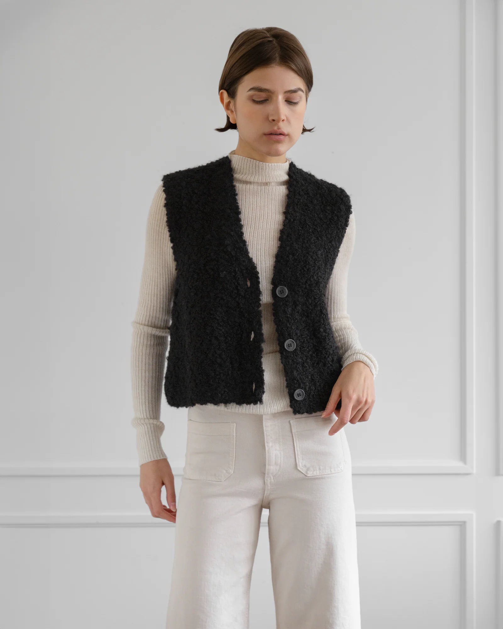 A person stands against a light gray wall wearing a cream ribbed turtleneck, high-waisted white pants, and the Aries Boucle Vest by LaTierra—a textured black alpaca blend vest with cow horn buttons.