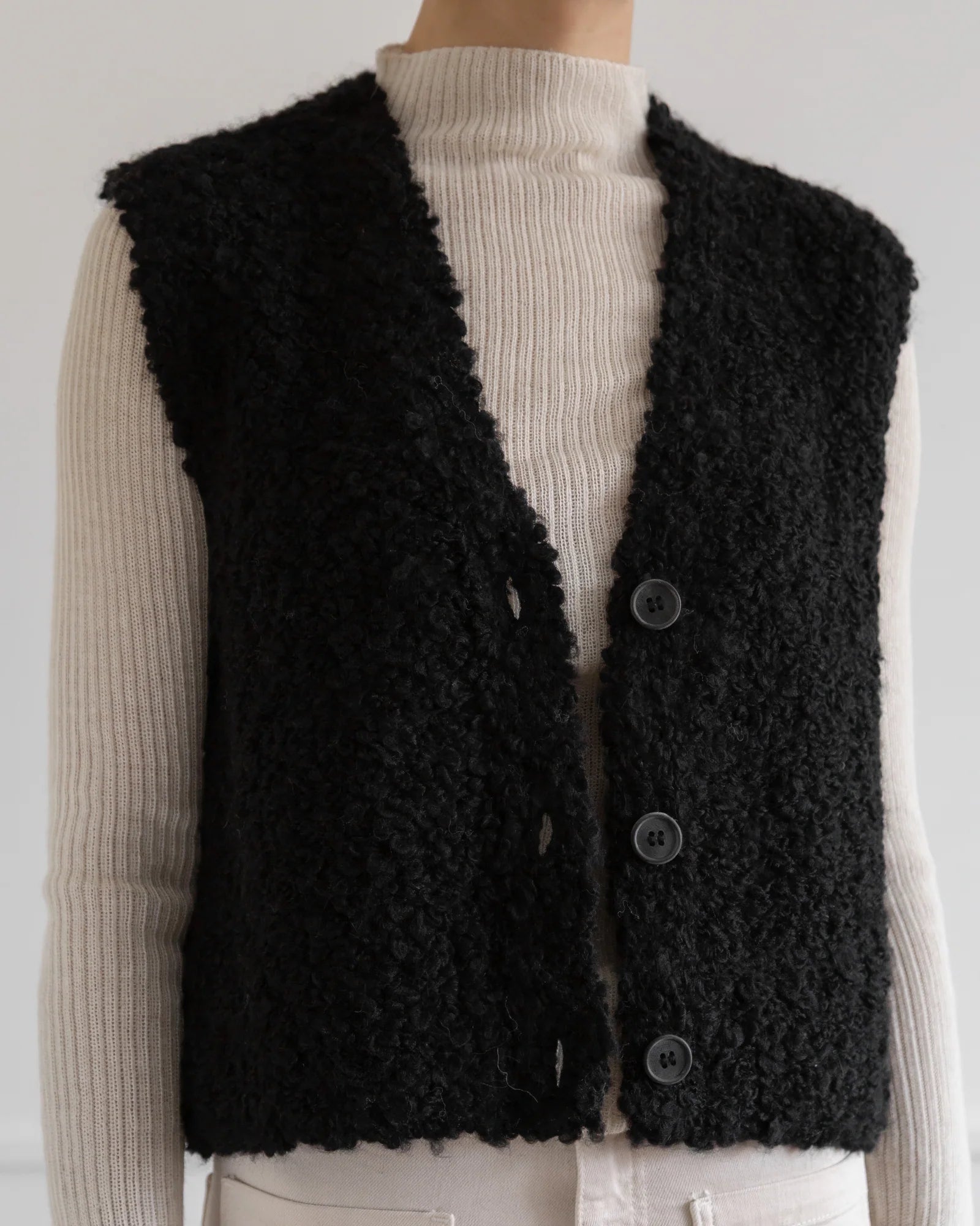 A person wears a cream ribbed turtleneck sweater with the LaTierra Aries Boucle Vest, a black, textured alpaca blend vest featuring a v neckline and three large cow horn buttons.