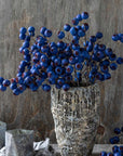Abigail Ahern’s Hawthorn Berry Midnight features vivid blue artificial berries arranged in a rustic vase, set against a textured wooden background. Two metallic cups complement the earthy, natural decor.