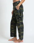 A person stands barefoot against a plain background, wearing ASKK's Carpenter pants in green camouflage. These stretch twill cargo pants, crafted with large side pockets and a relaxed fit, are made in Los Angeles. Their hands rest comfortably in the pockets.