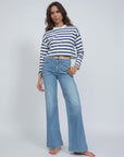 A person with long hair poses confidently in an ASKK Cropped LS Tee, a white and navy striped shirt made from American cotton, paired with blue bell-bottom jeans. They stand barefoot against a plain white background, exuding effortless style.