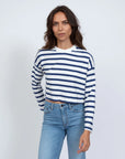 A person with long brown hair wears the ASKK Cropped LS Tee, featuring a white and blue stripe design. Paired with blue jeans, they stand against a plain white backdrop with a neutral expression, capturing the casual elegance of Los Angeles fashion.