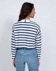 A person with long hair wears ASKK’s Cropped LS Tee, a blue and white striped long-sleeve top, paired with light blue jeans. Made in Los Angeles from American fabric, they stand away from the camera against a plain white background.