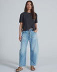 A woman with long brown hair and a relaxed expression stands against a plain background, wearing ASKK’s Cancun Pant—light blue Japanese denim jeans with wide legs and an elastic waistband—paired with a loose black t-shirt and tan sandals.