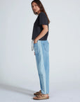A woman stands sideways with her hands in her pockets, wearing ASKK’s Cancun Pant—relaxed fit two-tone blue jeans with an elastic waistband—a black t-shirt, and tan sandals. Her long hair is down against a light gray background.