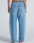 A person stands barefoot facing away, wearing light blue Cancun Pant jeans by ASKK with relaxed fit, back pockets, and an elastic waistband. The background is plain and light-colored.
