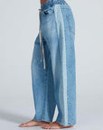 A person is wearing the ASKK Cancun Pant: light blue, wide-leg Japanese denim jeans with an elastic waistband, drawstring, and two-tone outer leg panels in a light Berkeley wash. The person is barefoot.