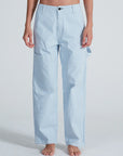 A person stands barefoot against a plain light background, wearing ASKK's Carpenter pants—light blue and white striped, high-waisted, wide-leg carpenter style with side pockets, crafted from stretch twill.