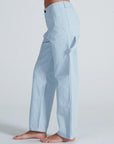 A person stands sideways barefoot against a plain background, wearing ASKK Carpenter relaxed fit light blue and white vertically striped stretch twill carpenter pants.