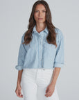 A woman with long brown hair wears the ASKK Cropped Nantucket Shirt—a blue and white vertically striped, lightweight cotton button-down—paired with high-waisted white pants against a plain light background.