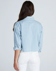 A woman with long brown hair, seen from behind, wears the ASKK Cropped Nantucket Shirt—blue and white striped Japanese shirting with double front pockets—paired with white pants, set against a light gray background.