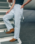A person in ASKK Carpenter light blue pinstriped pants, a dark sleeveless top, and brown boots stands on airplane stairs with one hand on their hip. The background shows the airplane and tarmac.