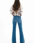 A woman with long wavy brown hair, wearing the ASKK Crosby Flannel in Japanese shirting fabric and high-waisted blue flared jeans, stands facing away against a plain white background, styled with black shoes.