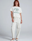A red-haired person wears the ASKK Shrunken Boy Tee in white with “TODAY'S SOUP IS A DIRTY MARTINI” text, cream pants, and pointed-toe shoes, standing in front of a plain gray background.