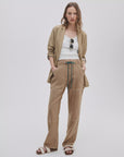 A woman poses with her hands in her tan Rag & Bone atlas linen pant, paired with a matching blazer and white tank. She accessorizes with sunglasses hanging from her shirt and completes the look with white sandals against a light backdrop.