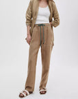 A person stands against a white background wearing a tan jacket over a white tank, Rag & Bone atlas linen pant in a relaxed fit, and beige buckled sandals; their face is not visible.