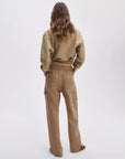 A person with wavy brown hair faces away, wearing a loose tan button-up shirt and relaxed fit high-waisted Rag & Bone atlas linen pant with visible back pockets, set against a plain light background.