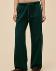 A person wears DayDreamer Drawstring Pants in dark green with a sunfaded effect and side pockets, styled with leopard print slippers, standing against a plain beige background.