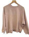 The Rehearsal Sweatshirt by The Great Inc.—a light pink, long-sleeve raglan sweatshirt—hangs on a wooden hanger against a white background, making it the perfect addition to your weekend essentials.