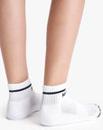 Close-up of a person’s lower legs and feet wearing Mother Baby Steps Ankle socks—classic black and white tube style with a black stripe and text—standing on a white surface.