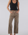 Wearing a black tank top and AMO Denim's Billie Pants in leopard print, a person stands confidently against a light background. The relaxed fit of the trousers is highlighted by their hands in the pockets, complemented by black loafers and long hair flowing freely.