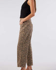 A person with long hair wears a black sleeveless top and AMO Denim's Billie Pants in leopard wash. Standing in side profile with hands in pockets and black loafers, the outfit's understated elegance contrasts beautifully with the plain white background.