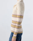 A person wearing the AMO Denim Desiree Tee, a cream-colored ribbed top with yellow, brown, and pink stripes, paired with blue jeans, stands sideways against a plain light background.