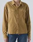 A person wearing the AMO Denim Jenn Shirt—a loose-fitting, tan corduroy top with front pockets and long sleeves—paired with blue jeans, stands against a plain light background.
