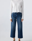 A person stands facing forward wearing a plain white long-sleeve shirt, AMO Denim Billie Pants—premium rigid denim cropped wide leg jeans—and black slip-on shoes against a light grey background.