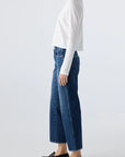 A person stands in profile wearing a long-sleeve white top, AMO Denim Billie Pants in premium rigid denim with a cropped wide leg, and black loafers against a plain light background.