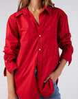 A person wearing AMO Denim's Ruth Oversized Shirt, bright red and vintage-inspired with sleeves rolled up, styled open-necked. Their hand is tucked into the pocket of stonewashed cotton poplin jeans. The face remains unseen.