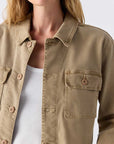 A woman models the Carly Jacket by AMO Denim—a light brown cotton lyocell piece—layered over a white ribbed crop top, shown from shoulders to waist against a plain background.