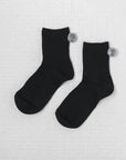 Unified's Winter Pom Pom Socks are cozy black ankle socks featuring small gray pompoms near the top cuffs, shown on a light textured surface.