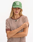 A woman wears the Clare Vivier Baseball Hat PasMal, featuring "PAS MAL" text and an adjustable metal closure, paired with a 100% cotton brown-and-white striped tee and layered necklaces, standing with arms crossed against a white background.