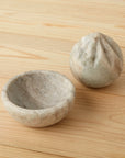 The Be Home Waterfall Marble Citrus Press, featuring a gray and white marble design with a ridged reamer and matching handmade bowl, sits on a light wood surface.