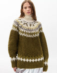A woman with long brown hair models the Beth Oversized Fair Isle Tneck by Rag & Bone, featuring an olive green and white pattern and high neck, standing against a plain white background.