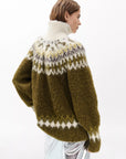A person with short brown hair wears the Rag & Bone beth oversized fair isle tneck, a chunky knit wool sweater in olive, white, yellow, and gray with a high collar, paired with a metallic silver skirt and turned slightly to the side.