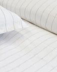Close-up of white bed sheets and pillowcases featuring the Blake Big Pillow 28x36 from POM POM, showcasing light gray interwoven stripes. The fabric appears soft and neatly arranged, creating a clean and minimalist look.