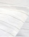 A close-up of the Blake Big Pillow 28x36 by POM POM reveals a white fabric adorned with subtle, sophisticated gray stripes, highlighting its hand-loomed texture and pattern. The material appears soft and lightweight, elegantly folded at an angle.