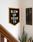 The Blow Up Your TV Pennant 18x24 by Oxford Pennant, a black flag with yellow trim and bold white text, hangs on a white wall near a painting of trees, above a potted plant and wooden staircase railing.
